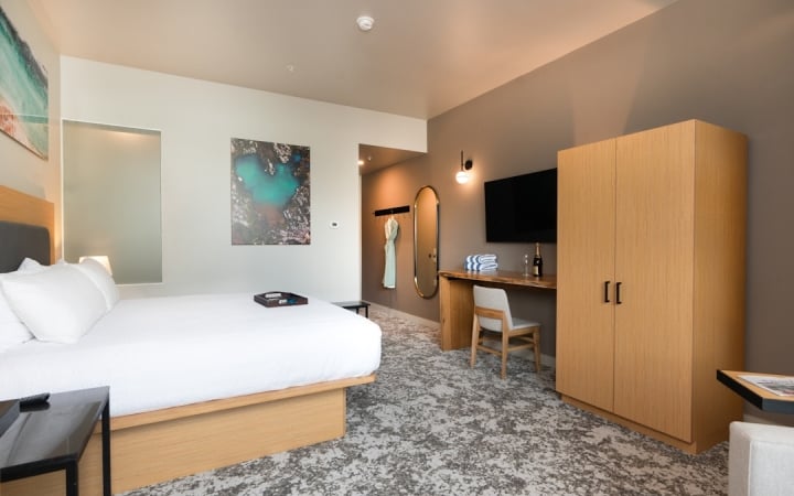 A made king-sized bed with white linens and a tray sits in a modern hotel room, which features a desk, wall-mounted TV, and light wood closet.