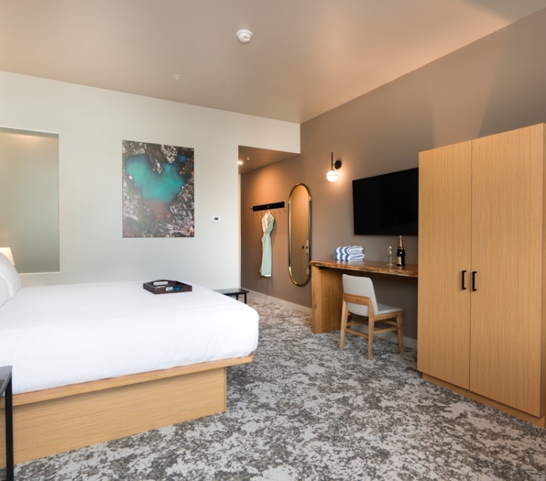 A made king-sized bed with white linens and a tray sits in a modern hotel room, which features a desk, wall-mounted TV, and light wood closet.