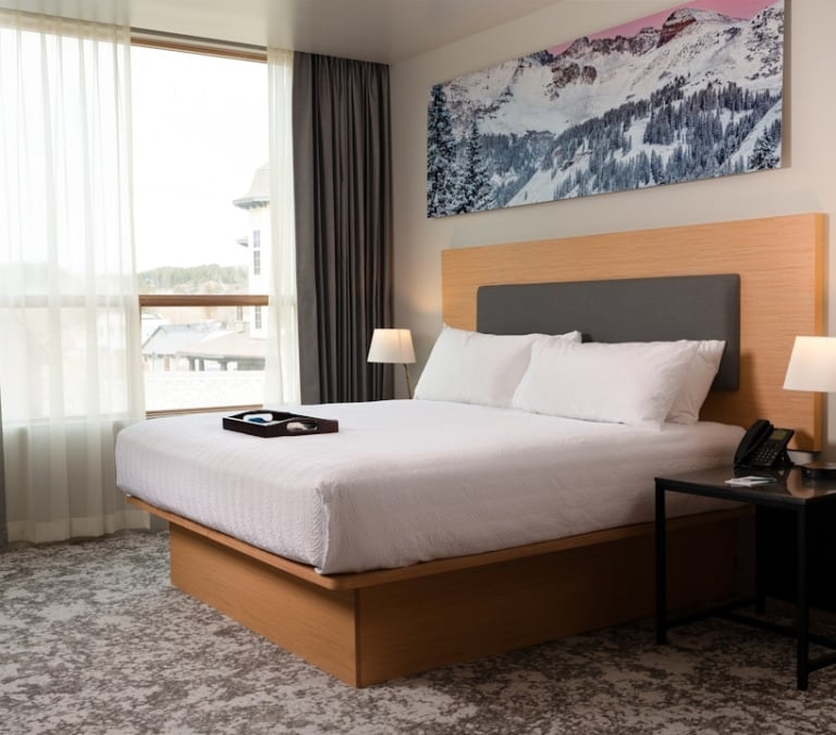 Queen bed with white linens and a tray in a hotel room. A large window with curtains overlooks a town, a snowy mountain painting above the bed.
