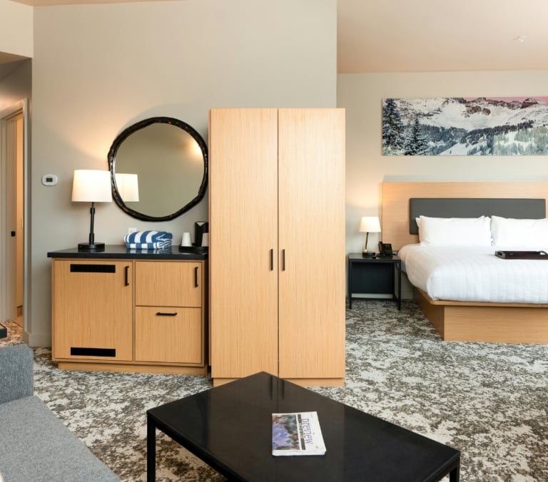 Modern hotel room furnished with a bed, sofa, and light wooden cabinetry on patterned carpet. A magazine titled 
