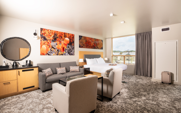 A modern hotel room with a queen bed, gray sofa, and two armchairs on patterned carpet. Large autumnal landscape art hangs above, a wide window overlooks buildings.