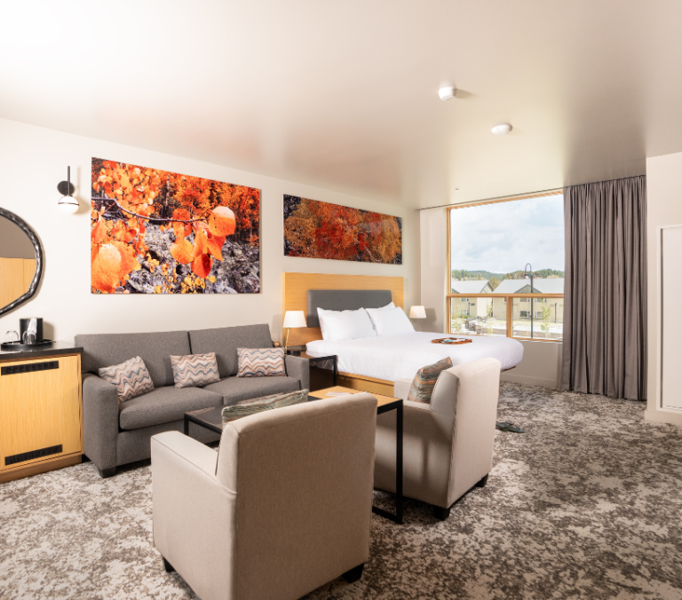 A modern hotel room with a queen bed, gray sofa, and two armchairs on patterned carpet. Large autumnal landscape art hangs above, a wide window overlooks buildings.