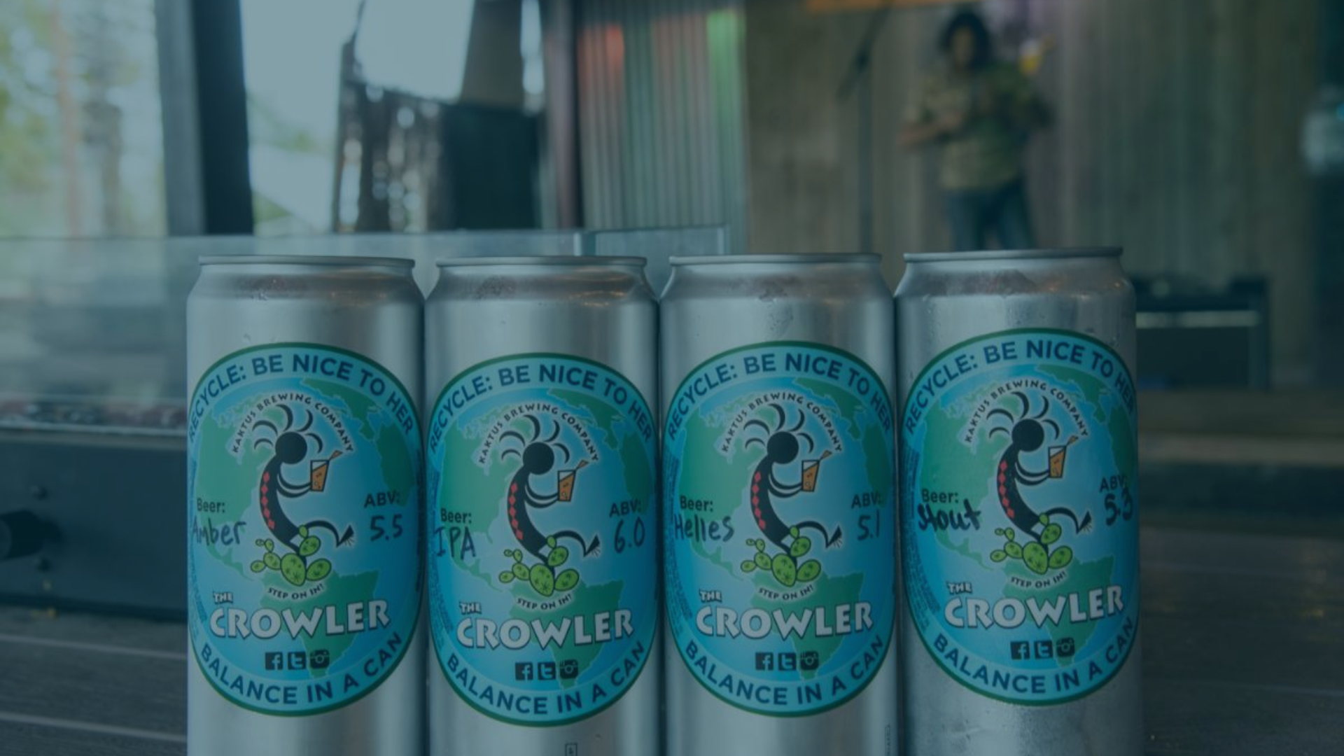 Four Kaktus Brewing Company beer cans stand in a row on a surface, with a blurred person on a stage behind them.
Text: RECYCLE: BE NICE TO HER, KAKTUS BREWING COMPANY, Beer: Amber ABV: 5.5, Beer: IPA ABV: 6.0, Beer: Helles ABV: 5.1, Beer: Stout ABV: 5.3, THE CROWLER, STEP ON IT!, BALANCE IN A CAN, f (Facebook icon), bird icon (Twitter), camera icon (Instagram).