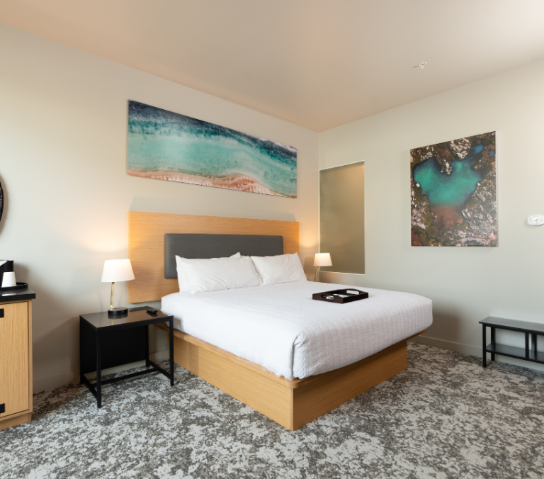 A modern queen bed, made with white linens and a tray, is centered in a hotel room. Walls feature abstract art, flanked by wooden furniture on patterned carpet.