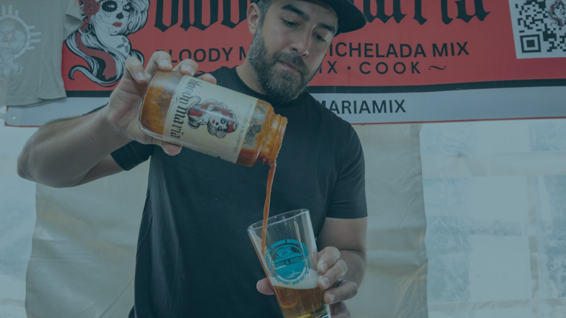A bearded man pours Bloody Maria mix from a jar into a beer glass, standing before a branded banner.
