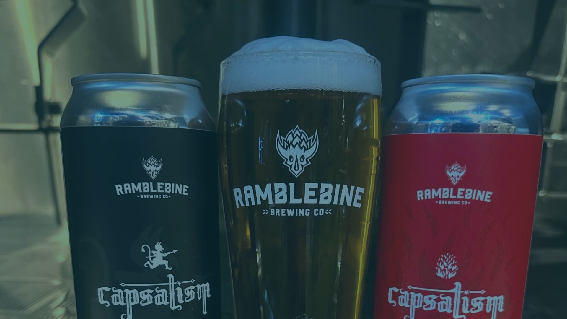 A frothy beer glass is centered between a black beer can and a red beer can, standing on a counter.
Text: RAMBLEBINE >> BREWING CO << (on all three items), capsalism (on both cans).