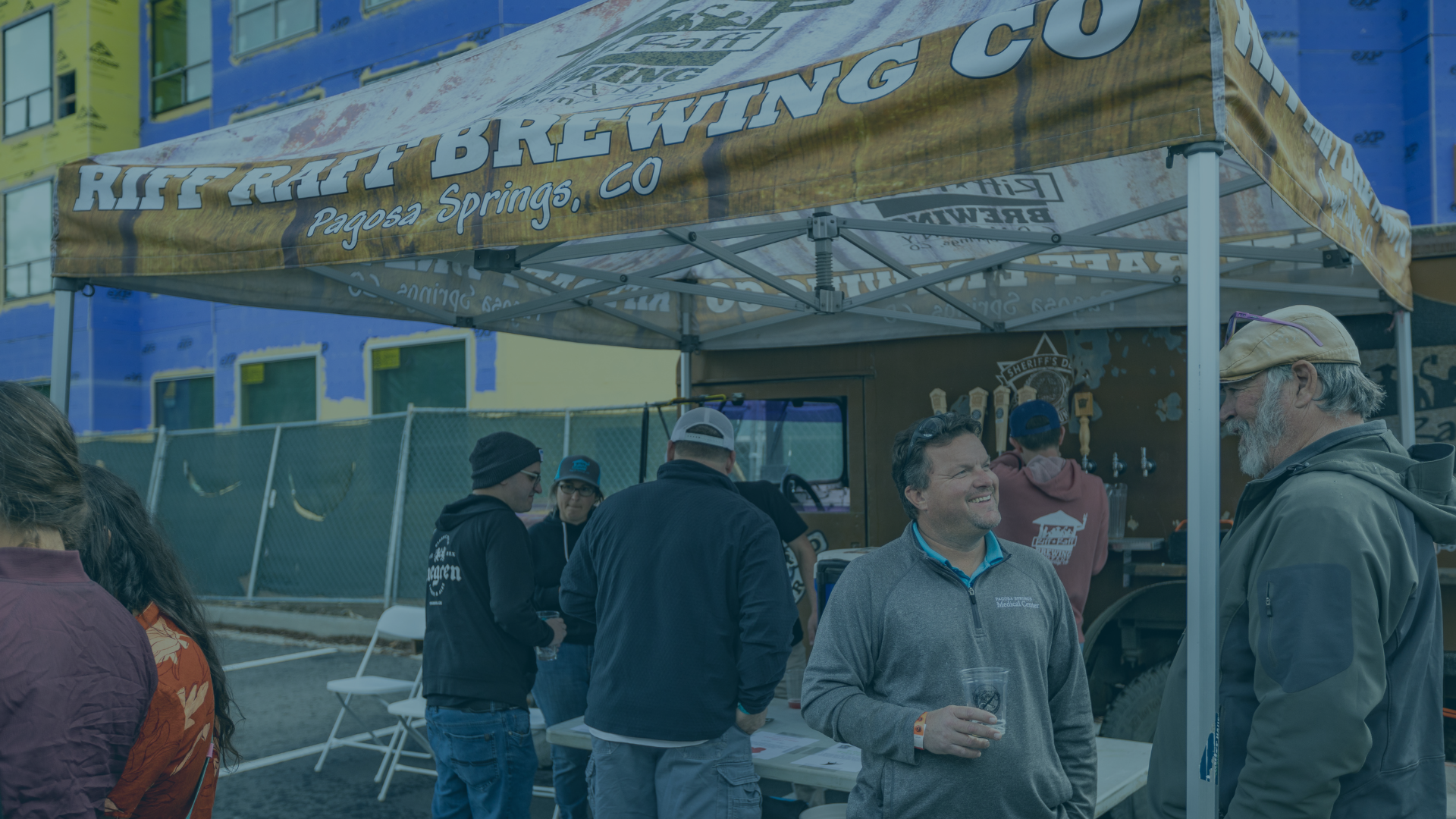 Several adults socialize, some holding cups, under a tent banner that reads: RIFF RAFF BREWING CO Pagosa Springs, CO. A building under construction is in the background. Other visible text: SHERIFF'S D, iregen, PAGOSA SPRINGS Medical Center, EXP.