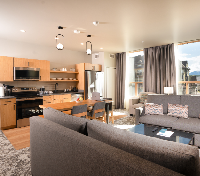 A modern open-plan apartment features a gray sofa facing a kitchen with light wood cabinets. Large windows reveal a village and snowy mountains outside. A bedroom is visible through an open door.