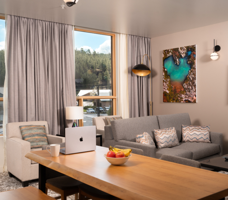 A laptop, fruit bowl, and mug rest on a wooden table in a modern hotel suite with a grey sofa, abstract art, and large windows overlooking a forested landscape.