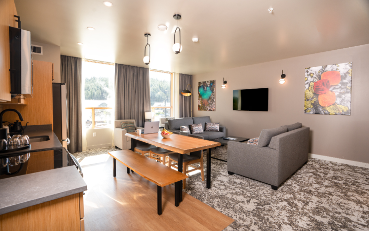 A modern, open-concept apartment features a kitchen, wooden dining table with a laptop, and a grey living room, all illuminated by large windows facing an outdoor landscape.