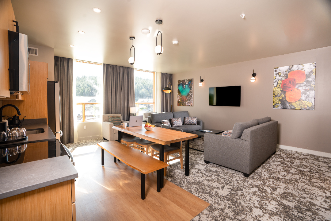 A modern, open-concept apartment features a kitchen, wooden dining table with a laptop, and a grey living room, all illuminated by large windows facing an outdoor landscape.