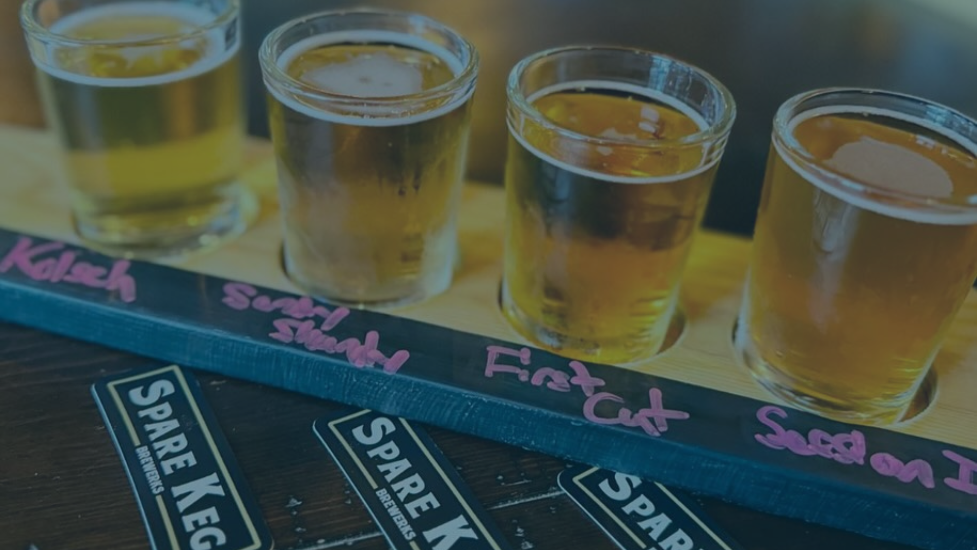 Four glasses of light-colored beer are presented in a wooden flight tray, labeled with chalk, alongside 