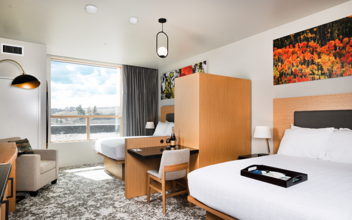 Modern hotel room features two beds, a desk with champagne, and a large window overlooking a hilly landscape under a blue sky.
