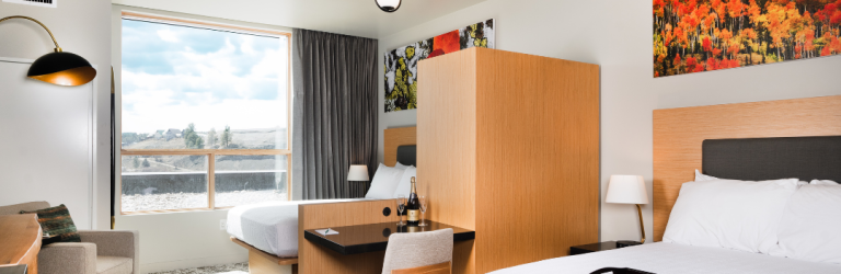 Modern hotel room features two beds, a desk with champagne, and a large window overlooking a hilly landscape under a blue sky.