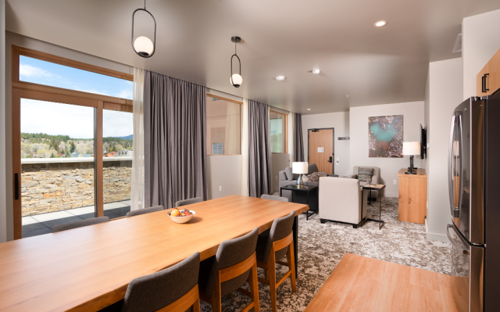 Long wooden dining table with fruit bowl and modern chairs in an open-plan room, extending to a living area and balcony with a town and mountain view.