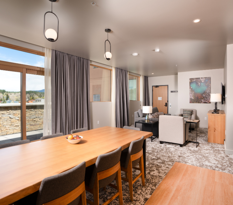 Long wooden dining table with fruit bowl and modern chairs in an open-plan room, extending to a living area and balcony with a town and mountain view.