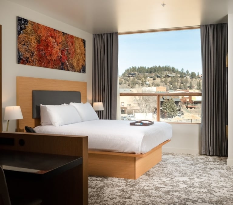 A modern hotel room features a large bed facing a window with a view of a hilly, tree-covered town. An abstract art piece hangs above the bed, with a desk area to the left.