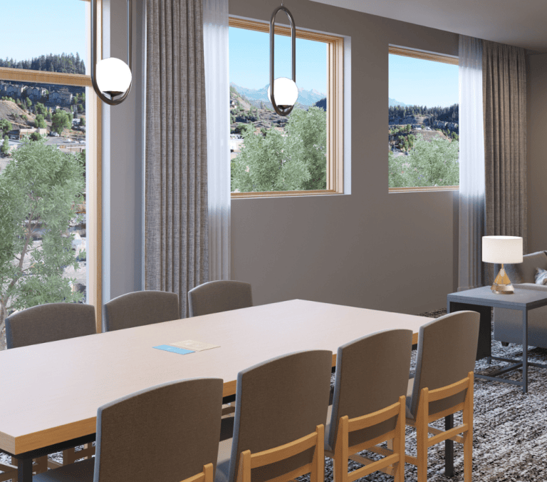 A spacious modern room features a long dining table with chairs and a lounge area. Large windows overlook a mountainous landscape with trees and distant buildings.