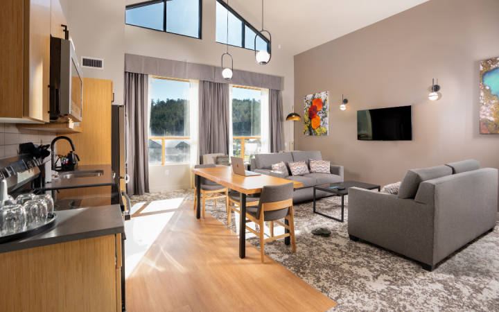 A modern open-plan kitchen and living room with light wood cabinetry, a dining table featuring a laptop, and two gray sofas. Large windows overlook trees.