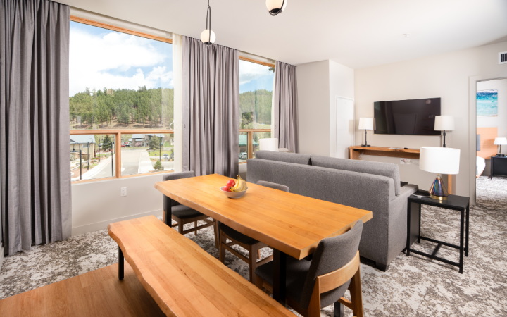 A modern dining area features a large wooden table with fruit, and a bench, next to a gray sofa and TV. Large windows overlook a town and forested hills.