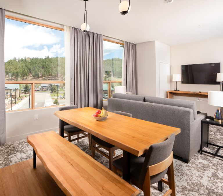 A modern dining area features a large wooden table with fruit, and a bench, next to a gray sofa and TV. Large windows overlook a town and forested hills.