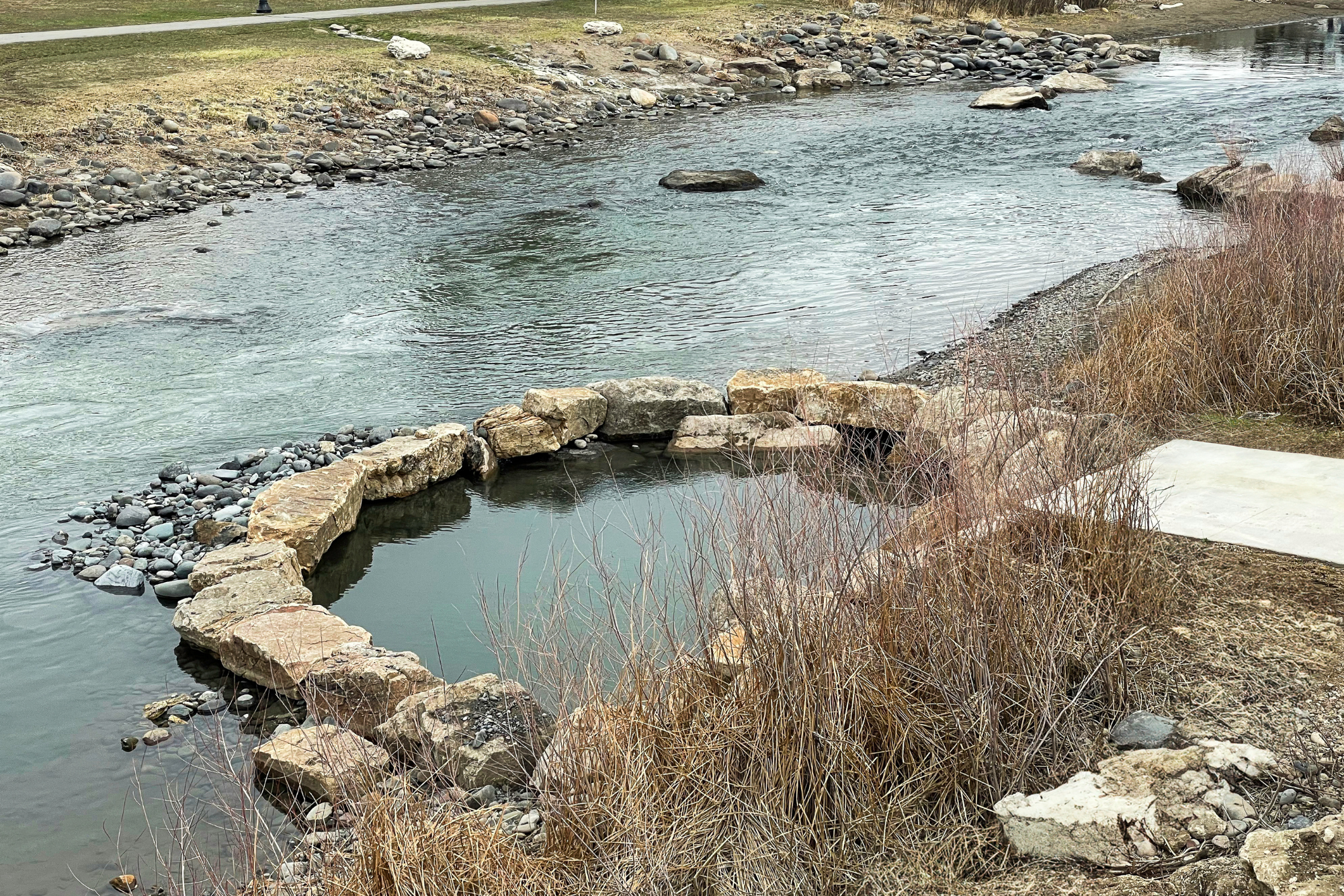 A circular stone-lined pool of still water sits on a rocky bank beside a flowing river. The far bank has grass and a path.