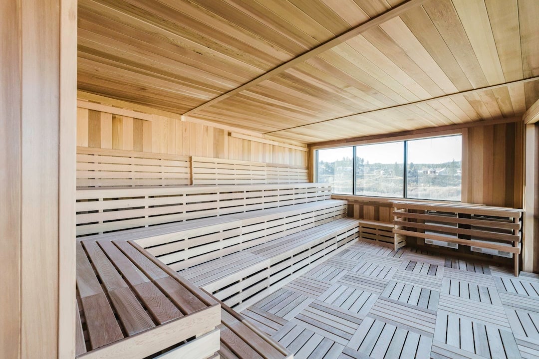 A wooden sauna room features tiered slatted benches along two walls and a wide window overlooking an outdoor landscape. The floor has slatted wood tiles.