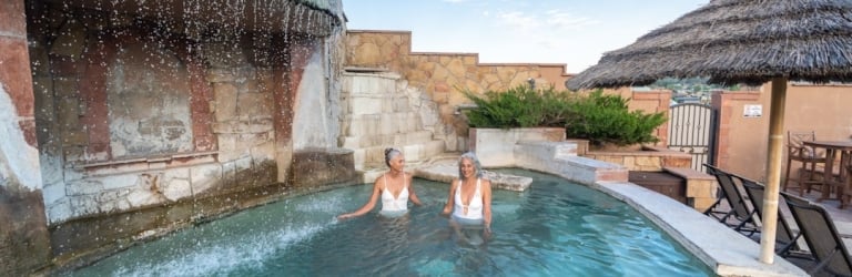 Two grey-haired women stand smiling in an outdoor pool, water cascading from a stone waterfall. The pool is part of a resort-like setting with a thatched umbrella under a blue sky.
Text: NO, NOTICE, NO LIFEGUARD ON DUTY, SWIM AT YOUR OWN RISK, NO DIVING, NO RUNNING, NO GLASS, NO ALCOHOL, NO PETS, NO SMOKING, THANK YOU.