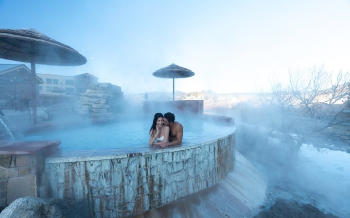 A man and woman are embracing in a steamy, blue outdoor hot spring. Stone-walled pools, thatched umbrellas, and resort buildings are visible under a clear sky.