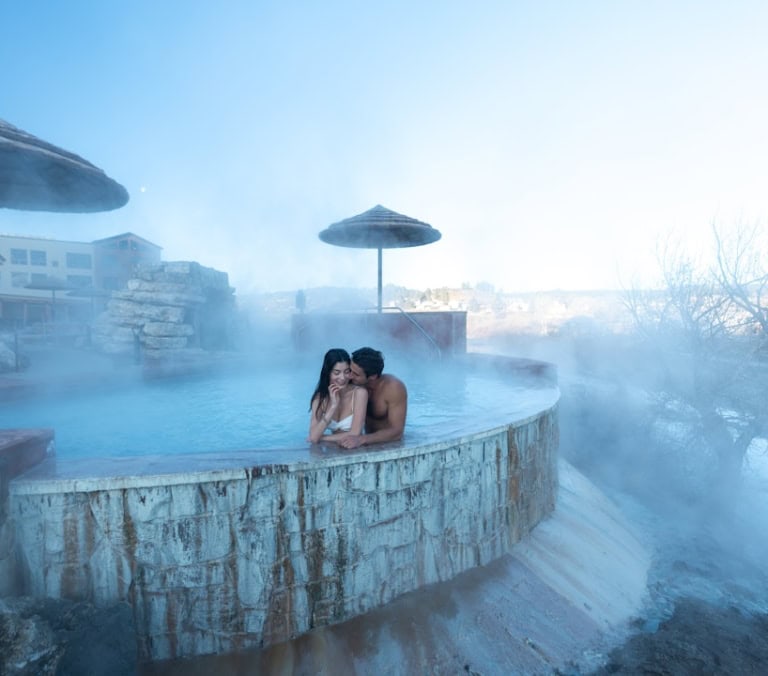 A man and woman are embracing in a steamy, blue outdoor hot spring. Stone-walled pools, thatched umbrellas, and resort buildings are visible under a clear sky.