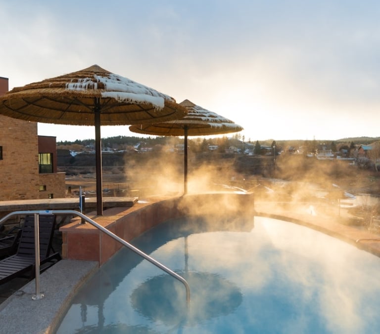 Steaming outdoor hot tub with a metal handrail, flanked by snow-covered thatched umbrellas, beside a brick building, overlooking a distant town at sunrise or sunset.