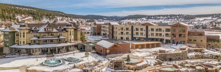 A large resort hotel and multiple steaming hot springs are nestled by a river in a snowy, mountainous landscape under a blue sky with clouds.