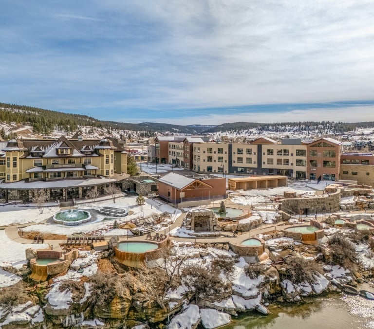 Numerous geothermal pools cascade towards a river, bordered by a large hotel (S SPRINGS) and multi-story buildings (SPA) against snow-dusted mountains.
