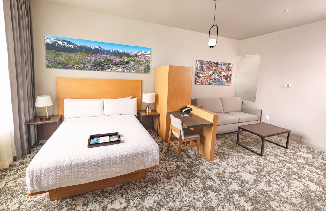 A contemporary hotel room is furnished with a white-linened bed, a desk area with a chair, and a beige sofa, all set on patterned carpet with various artworks on the walls.