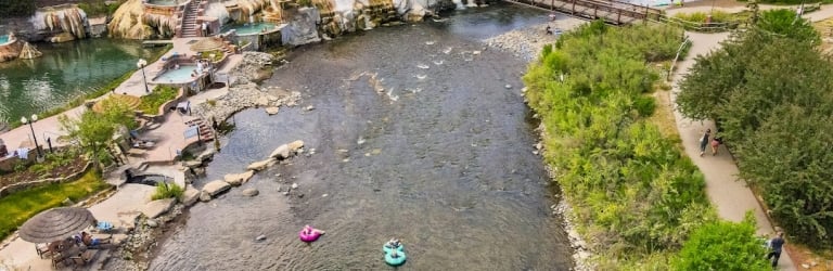 River flows past terraced hot springs with people soaking, and a tree-lined path. A large hotel (CAFE visible) overlooks this mountain town scene.