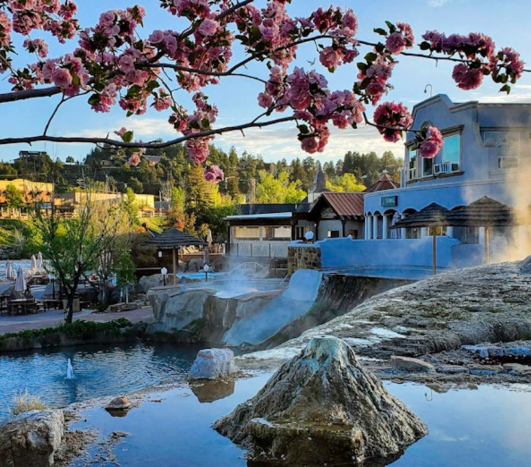 Steaming mineral springs cascade into tranquil pools, framed by pink cherry blossoms. A blue SPA resort building and a distant town are visible under a clear sky.