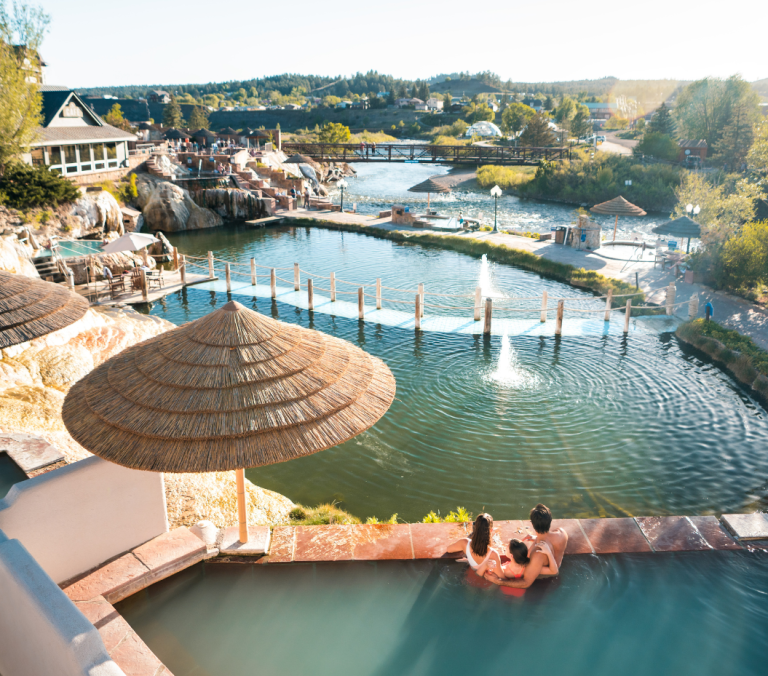 A family relaxes in an outdoor hot spring pool, overlooking a winding river with fountains, straw umbrellas, and resort buildings. Bridge text: PAGOSA RIVERWALK.