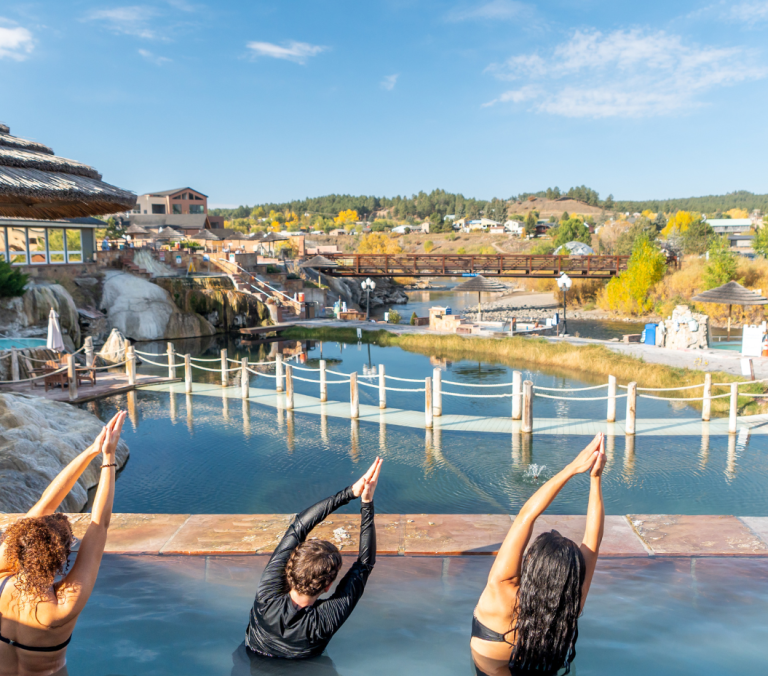 Four people in a hot spring pool practice yoga with raised arms, overlooking a river, bridges, and a town with vibrant fall foliage under a clear blue sky.