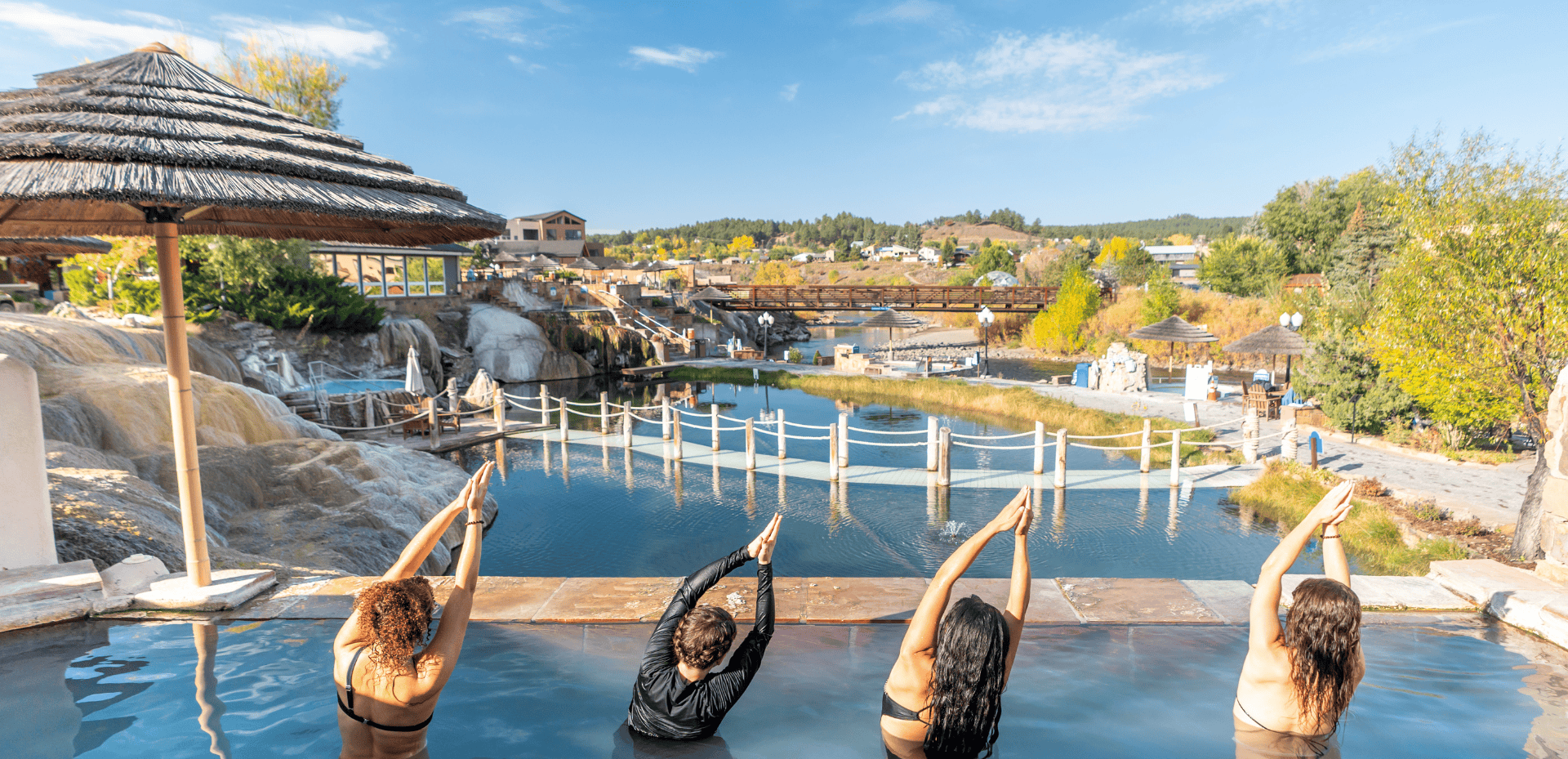 Four people in a hot spring pool practice yoga with raised arms, overlooking a river, bridges, and a town with vibrant fall foliage under a clear blue sky.