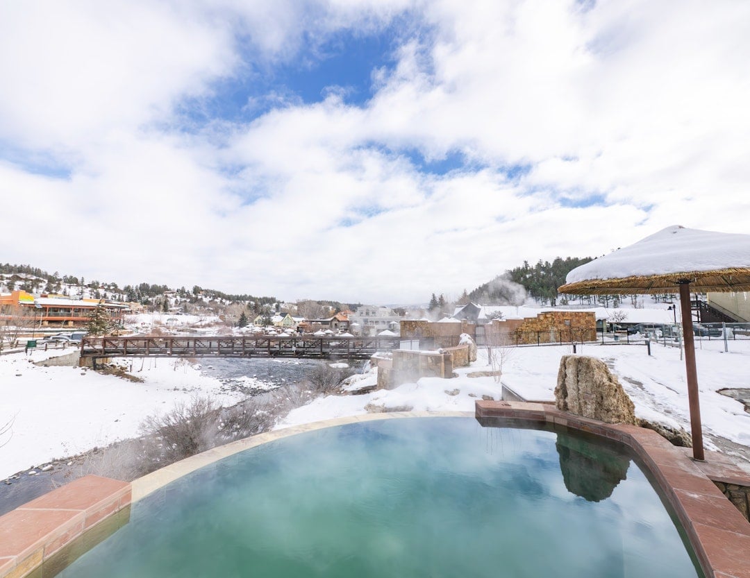 Steaming, green thermal pool reflects a cloudy blue sky, surrounded by snow, overlooking a river, a bridge, and a distant snowy town.