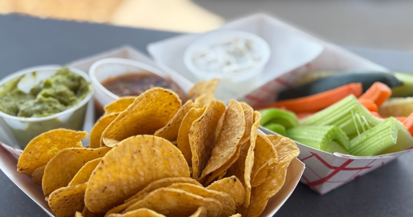 Crisp tortilla chips fill a paper tray, accompanied by cups of guacamole, salsa, creamy dip, and a tray of cut celery and carrots on a dark surface.