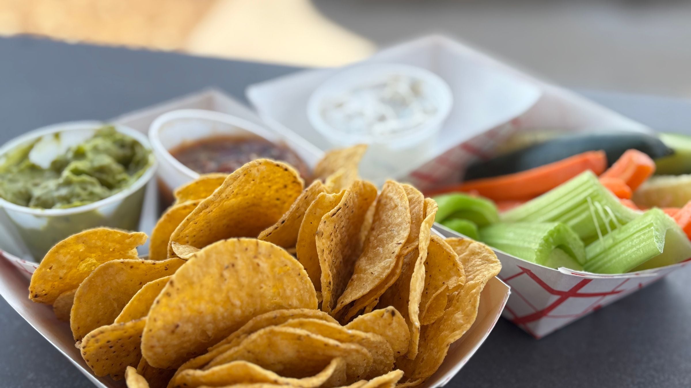 Crisp tortilla chips fill a paper tray, accompanied by cups of guacamole, salsa, creamy dip, and a tray of cut celery and carrots on a dark surface.