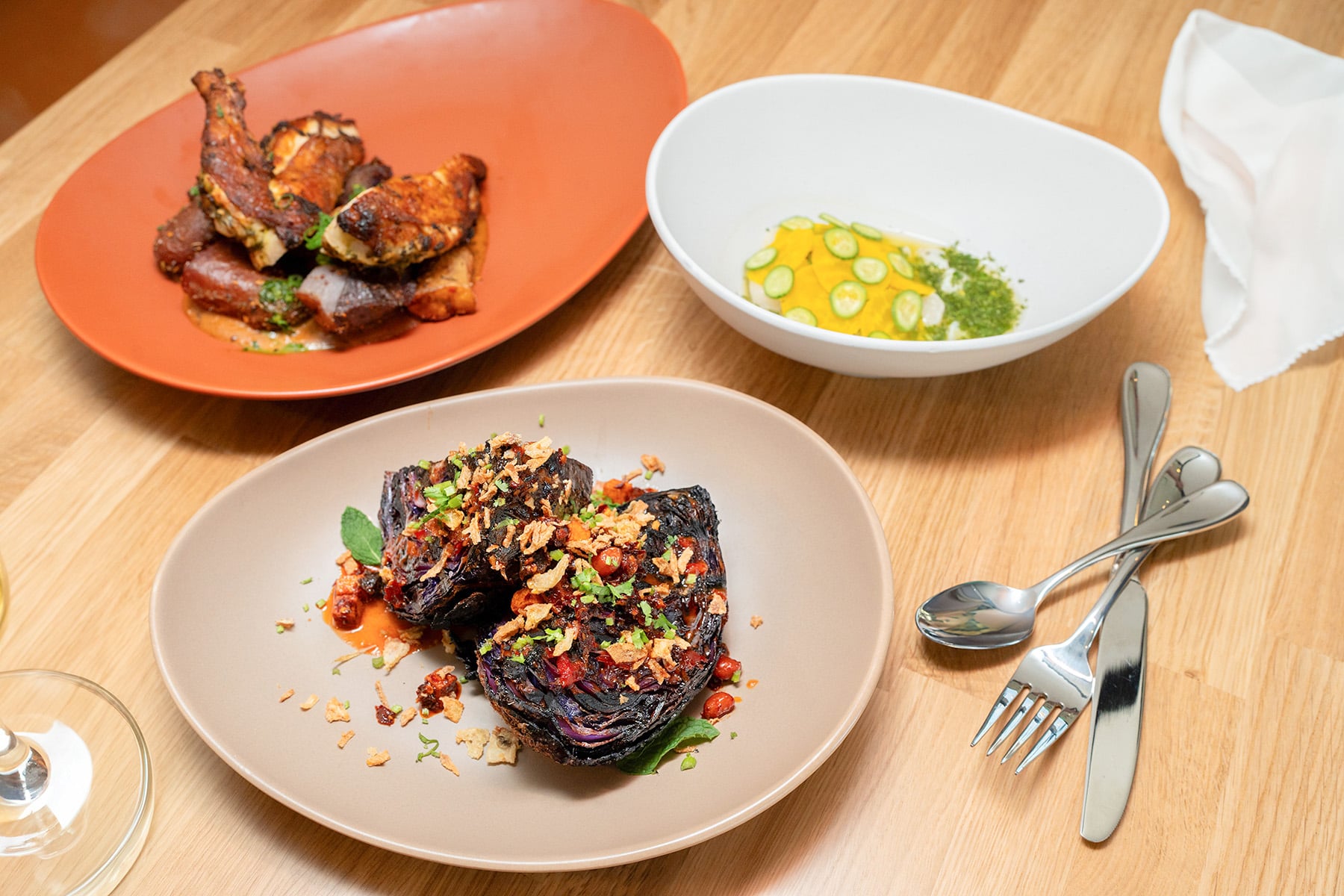 Three gourmet dishes: grilled meat, charred cabbage, and a yellow puree with cucumber, presented on plates and a bowl on a light wooden table with cutlery.