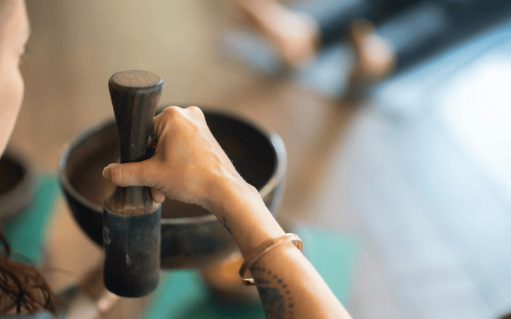 A woman's hand grips a wooden mallet, poised above a brass singing bowl in a blurred indoor setting, suggesting meditation or sound healing.