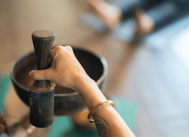 A woman's hand grips a wooden mallet, poised above a brass singing bowl in a blurred indoor setting, suggesting meditation or sound healing.