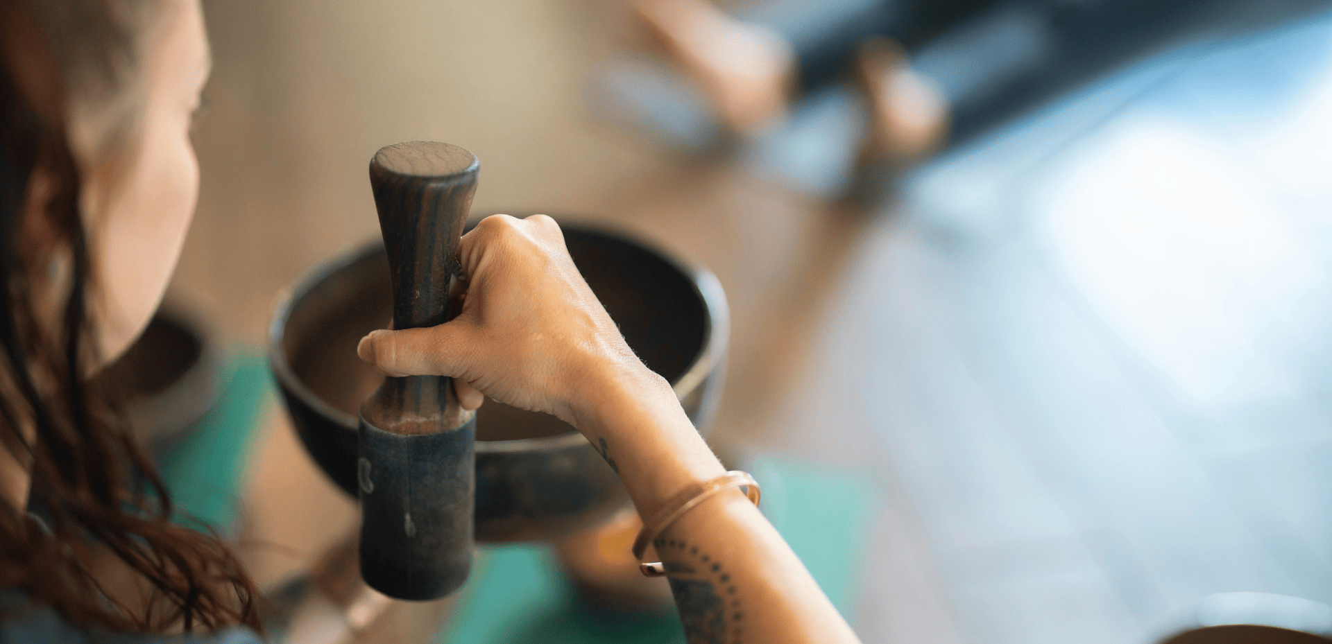 A woman's hand grips a wooden mallet, poised above a brass singing bowl in a blurred indoor setting, suggesting meditation or sound healing.