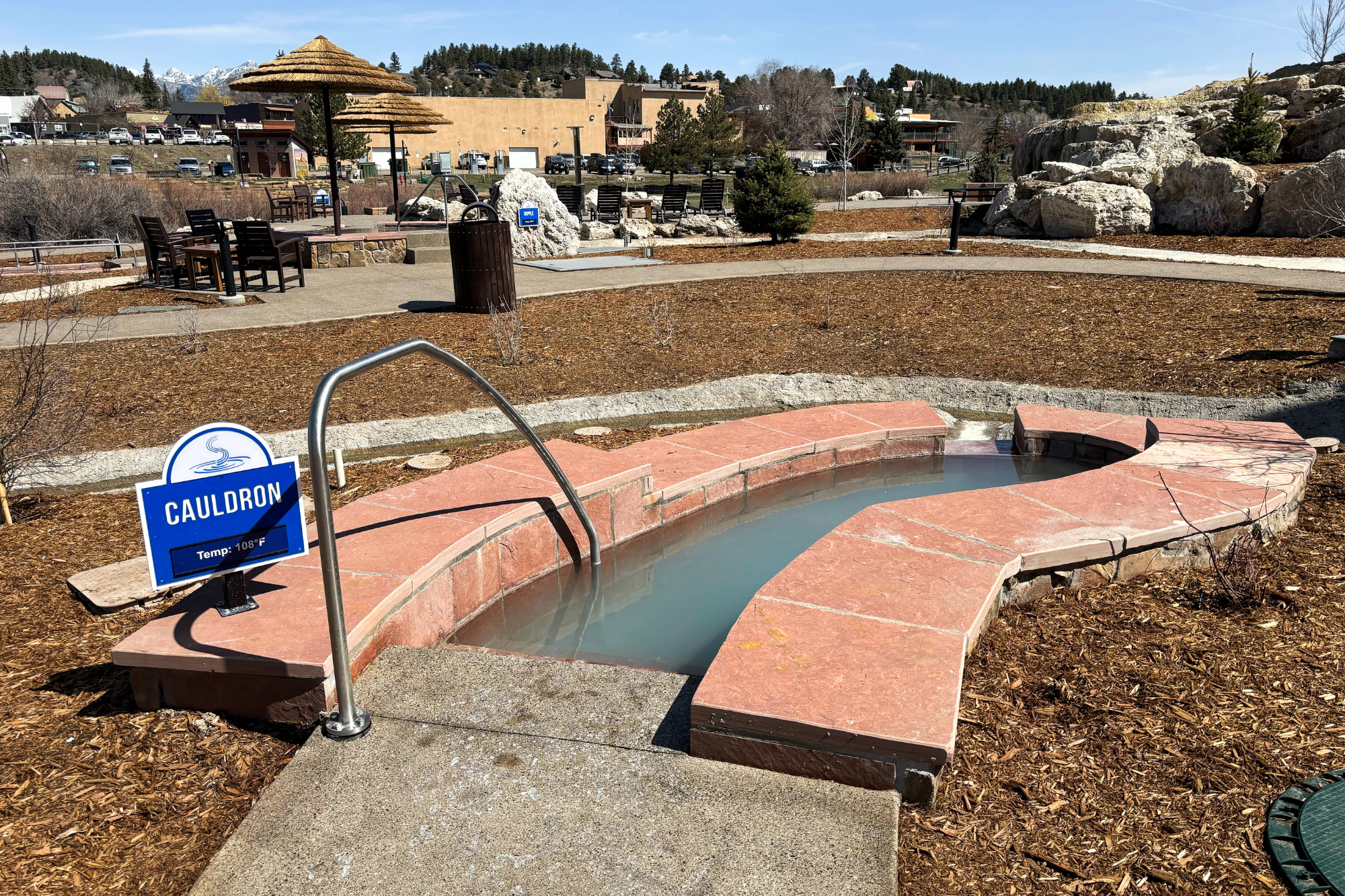 A curved, red-tiled hot spring pool filled with pale grey water and a metal handrail, located in a mulched outdoor area with distant town buildings and mountains. Text: CAULDRON, Temp: 108°F