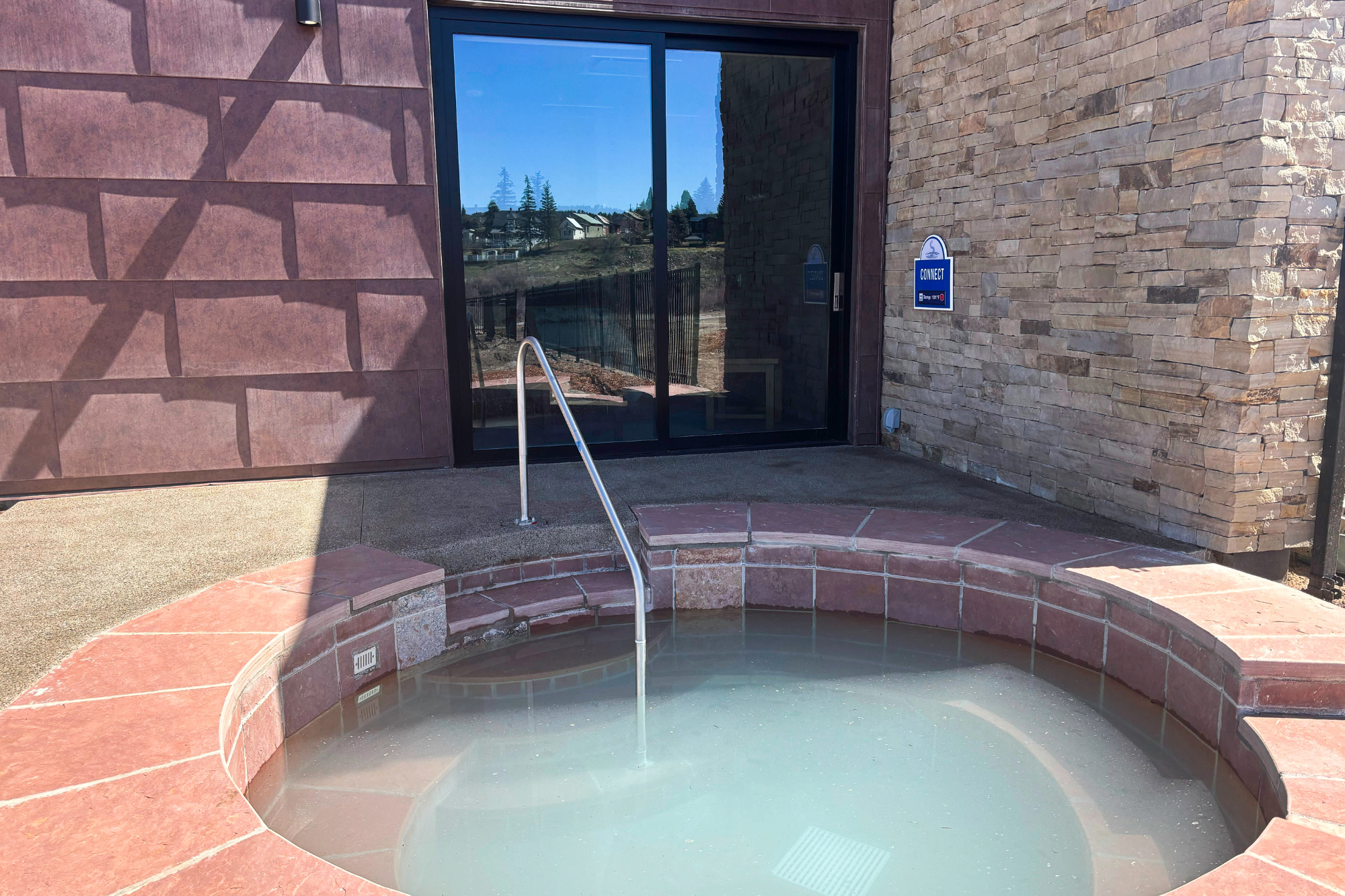 An outdoor, red-tiled hot tub filled with light blue water and a metal handrail sits next to a building. A blue sign on the wall reads: 