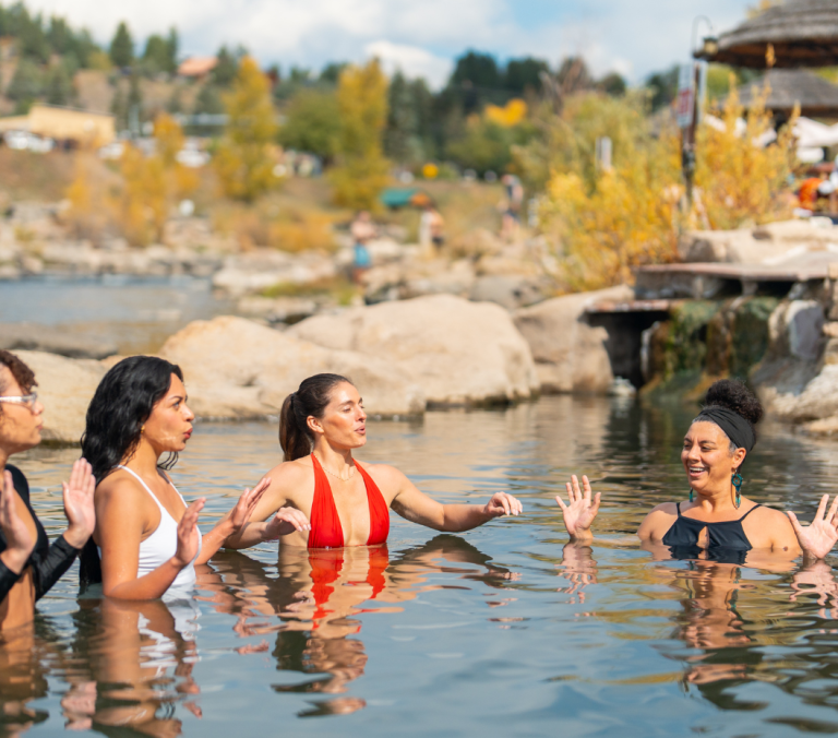 Four women are chest-deep in a natural-looking pool, gesturing and interacting. The background shows rocks, a small waterfall, and a distant village with autumn foliage.
