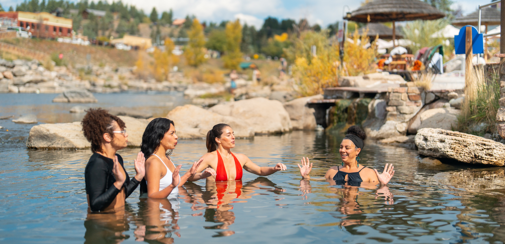 Four women are chest-deep in a natural-looking pool, gesturing and interacting. The background shows rocks, a small waterfall, and a distant village with autumn foliage.